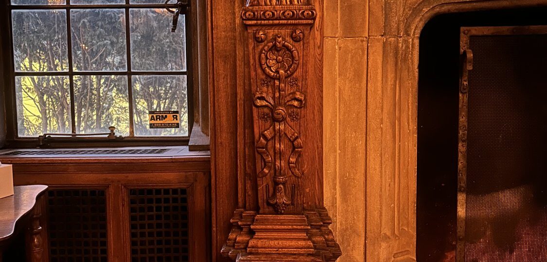 The carved stone chimneypiece in the library of the Pillsbury Mansion, flanked by carved oak figures and lit by leaded glass, bearing the Tallow Chandlers guild arms granted in 1456.