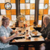 Two guests seated at a table inside the Pillsbury Club with food and drinks in front of them and a wall of amber and orange stained glass behind.