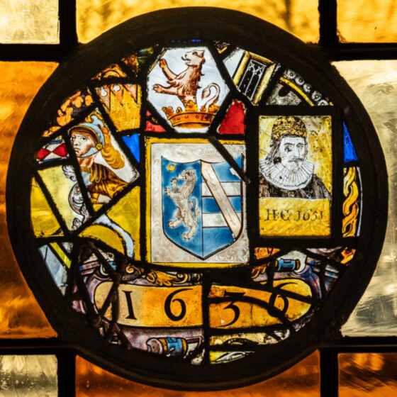 A leaded stained glass roundel in a Pillsbury Mansion window: a heraldic shield, a crowned lion, a helmeted figure, a bearded man in a ruff and the date 1636.